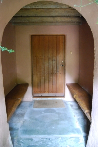 A long narrow porch, entered through an arch with thick plaster walls, painted pink. Inside, the floor is made of large stone slabs; a wooden plank along each  side wall form benches. Timber beams run across the ceiling; and at the end is a wooden door.