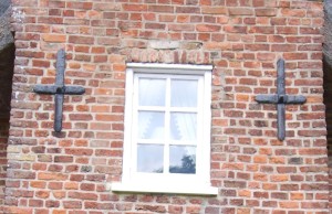 A narrow section of an old house wall, wall, with a small, central window, and a large, iron, cross either side, to stabilise the wall.