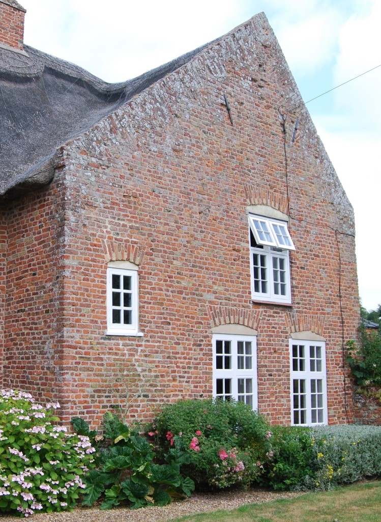 A projecting wing of an old building, constructed of small, irregular, red bricks, and topped by a thatched roof. The gable-wall contains two ground floor windows, one first floor window (placed between the two below), and one at a level between the two floors.